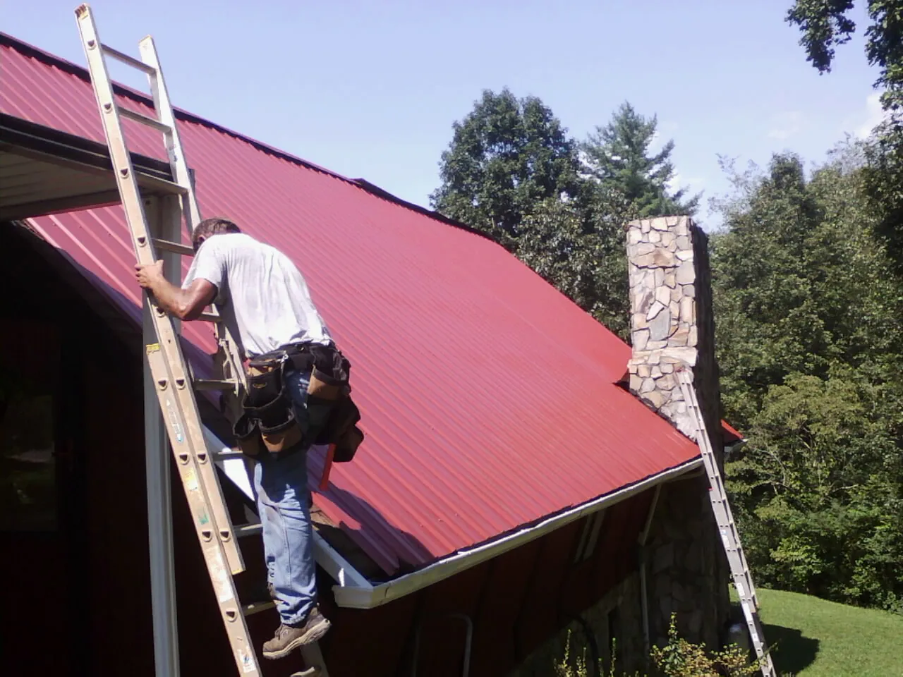 Roofer on a ladder beside a red metal roof and stone chimney — Soffit & Fascia Repair by Roofing Brothers in Fenwick Island